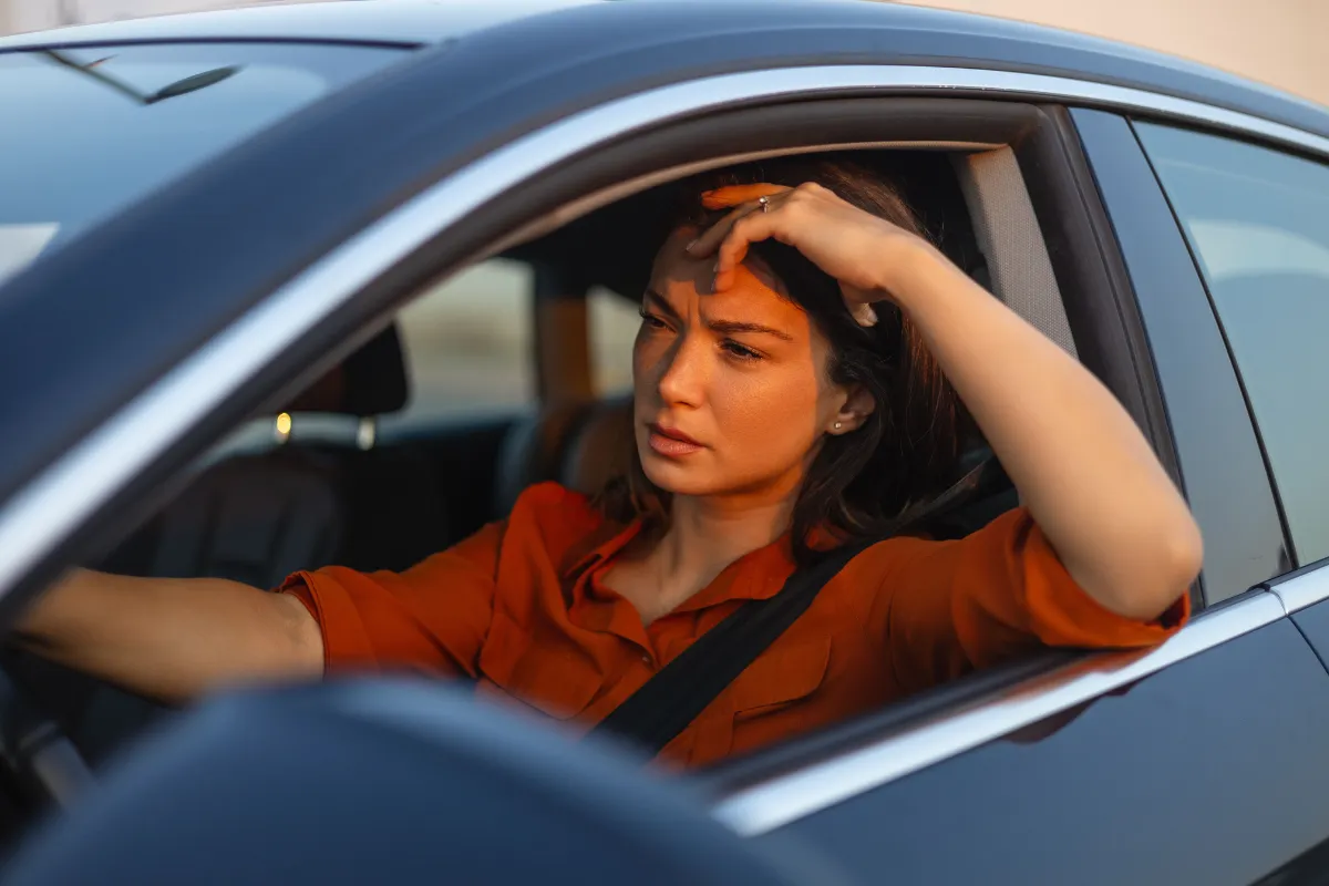Mulher com a mão na cabeça sofrendo com exposição ao sol dentro do carro com a janela aberta